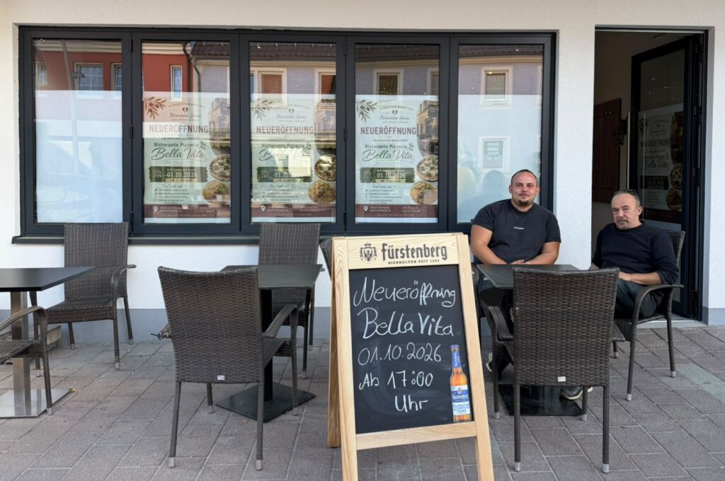 Francesco und Giuseppe mit Schild Neueröffnung am Tisch vor ihrem Laden